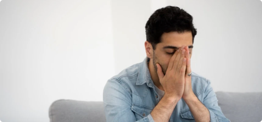 En mann sitter på en sofa med hendene som dekker ansiktet, og viser tegn på fortvilelse eller angst. Han ser ut til å være overveldet av følelsene sine.